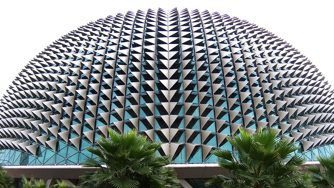 Fachada de domo geométrico con paneles metálicos triangulares repetitivos que crean un llamativo patrón de chevrones, con palmeras visibles en la base de la estructura arquitectónica moderna